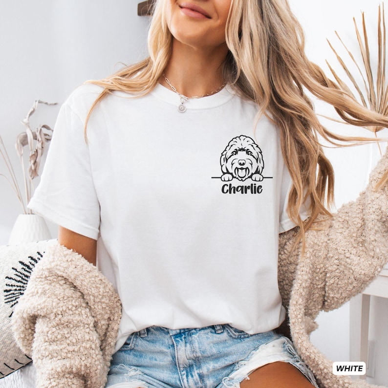 Op de afbeelding: Een wit T-shirt met korte mouwen voorzien van een zwarte lijntekening van een hondenhoofd dat boven een horizontale lijn uitsteekt, met de naam Charlie in een vetgedrukt zwart lettertype direct onder de hondenillustratie op de linkerborst.