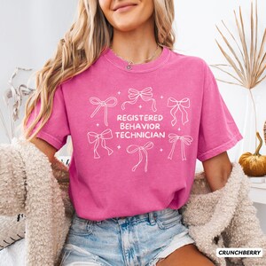 May include: A bright pink short-sleeve t-shirt featuring white line art of six bows arranged around the text "REGISTERED BEHAVIOR TECHNICIAN" in a bold, rounded font. The shirt has a crew neckline and a relaxed fit, with small star accents.
