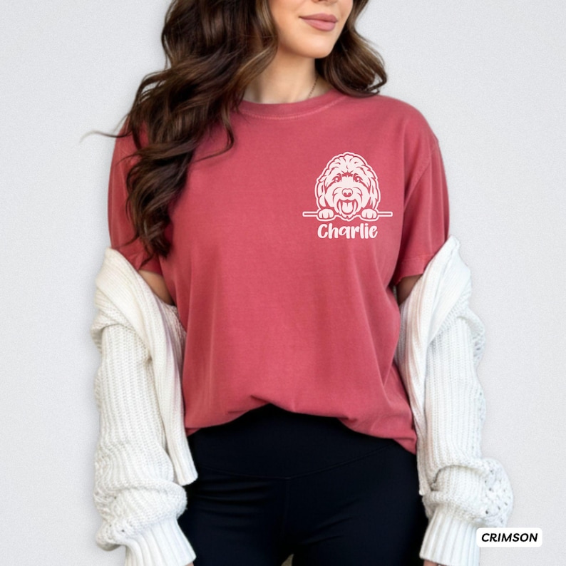 Op de afbeelding: Een karmozijnrood T-shirt met korte mouwen voorzien van een witte lijntekening van een hond die over een horizontale lijn kijkt, met de naam Charlie in wit schrift onder de hond gedrukt op de linkerborst van het kledingstuk.