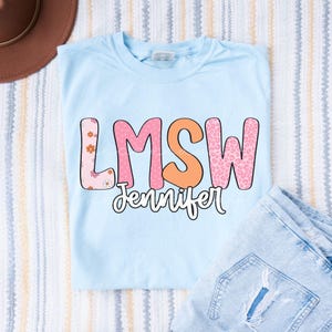 Puede incluir: Una camiseta azul claro con las letras LMSW en una fuente grande y estampada con diseños de flores rosas, rosa sólido, naranja sólido y estampado de leopardo rosa. Debajo de las letras, el nombre Jennifer está impreso en una fuente cursiva blanca.