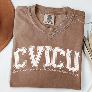 May include: A brown t-shirt with the text "CVICU Cardiovascular Intensive Care Unit" printed in white. The shirt has a distressed look and is made of a soft, comfortable material.