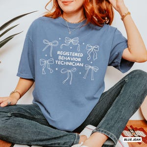 May include: A blue short-sleeved t-shirt featuring white line art of six bows and the text "REGISTERED BEHAVIOR TECHNICIAN" printed in the centre. The shirt is paired with dark blue denim jeans, creating a casual look with a graphic design.