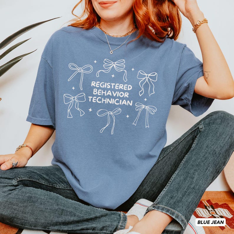 May include: A blue short-sleeved t-shirt featuring white line art of six bows and the text "REGISTERED BEHAVIOR TECHNICIAN" printed in the centre. The shirt is paired with dark blue denim jeans, creating a casual look with a graphic design.