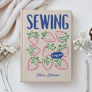 May include: A beige journal with the word "SEWING" in blue, and illustrations of strawberries. The phrase "is my jam" is in a blue oval, and the name "Olivia Johnson" is below. The journal is surrounded by white flowers and a pen.
