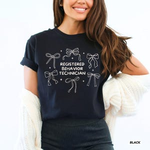 May include: A black short-sleeved t-shirt featuring a white graphic print of five decorative bows arranged around the text "REGISTERED BEHAVIOR TECHNICIAN" in a clean, sans-serif font, accented by small four-pointed star icons.
