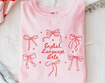 May include: A light pink crewneck t-shirt featuring a red graphic print of five various ribbon bows and small four-pointed stars surrounding the centered red text that reads English Language Arts in a cursive script font.