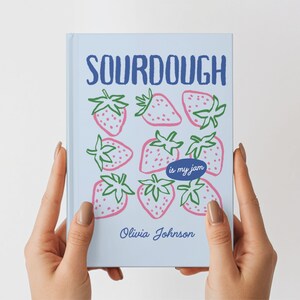 Puede incluir: Un diario azul claro con la palabra "SOURDOUGH" en azul oscuro e ilustraciones de fresas rosas con tallos verdes. La frase "is my jam" est&aacute; en un &oacute;valo azul, y el nombre "Olivia Johnson" est&aacute; en la parte inferior.