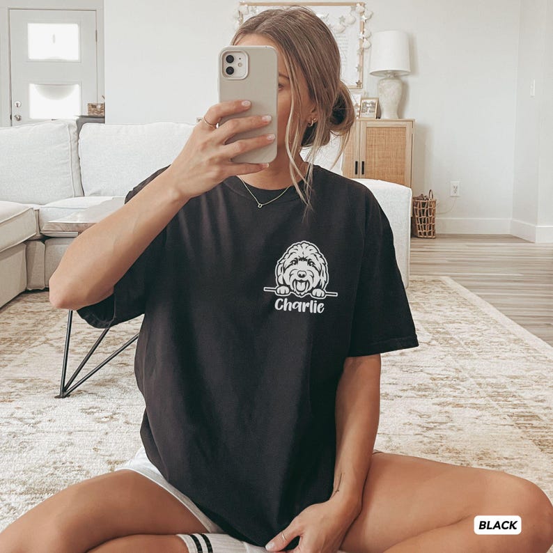 Op de afbeelding: Een zwart t-shirt met korte mouwen en een witte afbeelding van een hond die over een horizontale lijn kijkt, met de naam Charlie eronder gedrukt in een cursief lettertype. Het shirt heeft een ronde hals en een relaxte pasvorm.