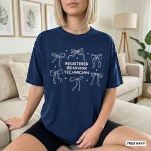 May include: A navy blue short-sleeved t-shirt featuring white line art of six decorative bows surrounding the centred text "REGISTERED BEHAVIOR TECHNICIAN" in a clean, sans-serif font. The garment has a crew neckline and a relaxed fit.