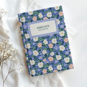 May include: A hardcover journal with a floral pattern in blue, white, and pink. The cover features the text "AMELIA'S NEW CHAPTER" in a decorative frame. The journal is set on a white cloth background.