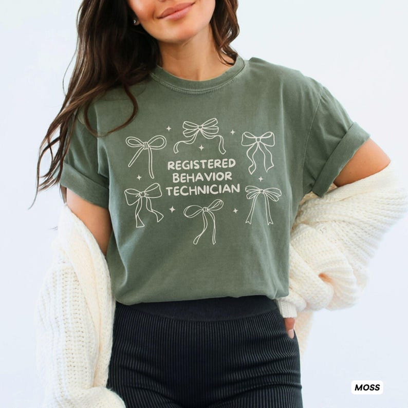 May include: A moss green short-sleeve t-shirt featuring a white graphic print of six decorative bows and small four-pointed stars surrounding the centred text "REGISTERED BEHAVIOR TECHNICIAN" in a clean, sans-serif, uppercase font style.