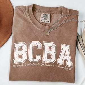 May include: A brown t-shirt with the text "BCBA Board Certified Behavior Analyst" printed in white. The shirt is made of a soft, comfortable material.