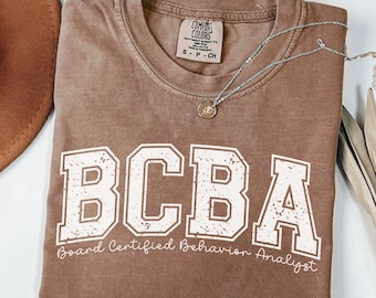 May include: A brown short-sleeve t-shirt with a crew neckline and a Comfort Colors brand label. The front features large white block letters reading BCBA and the text Board Certified Behavior Analyst in a script font below the letters.