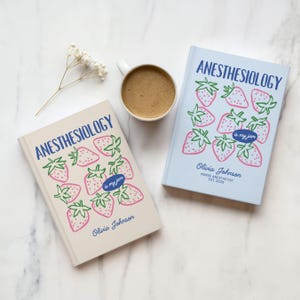 May include: Two hardcover books titled Anesthesiology is my jam with strawberry illustrations, one in cream and one in light blue. Each cover features the name Olivia Johnson and a coffee mug with brown liquid sits between the two books.