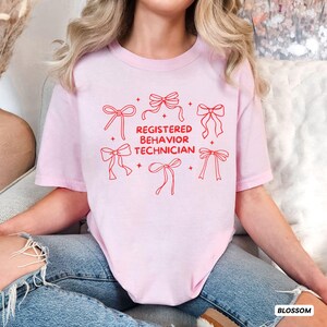 May include: A light pink short-sleeved t-shirt featuring red line art graphics of six bows arranged around the centred red text that reads REGISTERED BEHAVIOR TECHNICIAN, accented by small four-pointed star shapes scattered throughout the design.