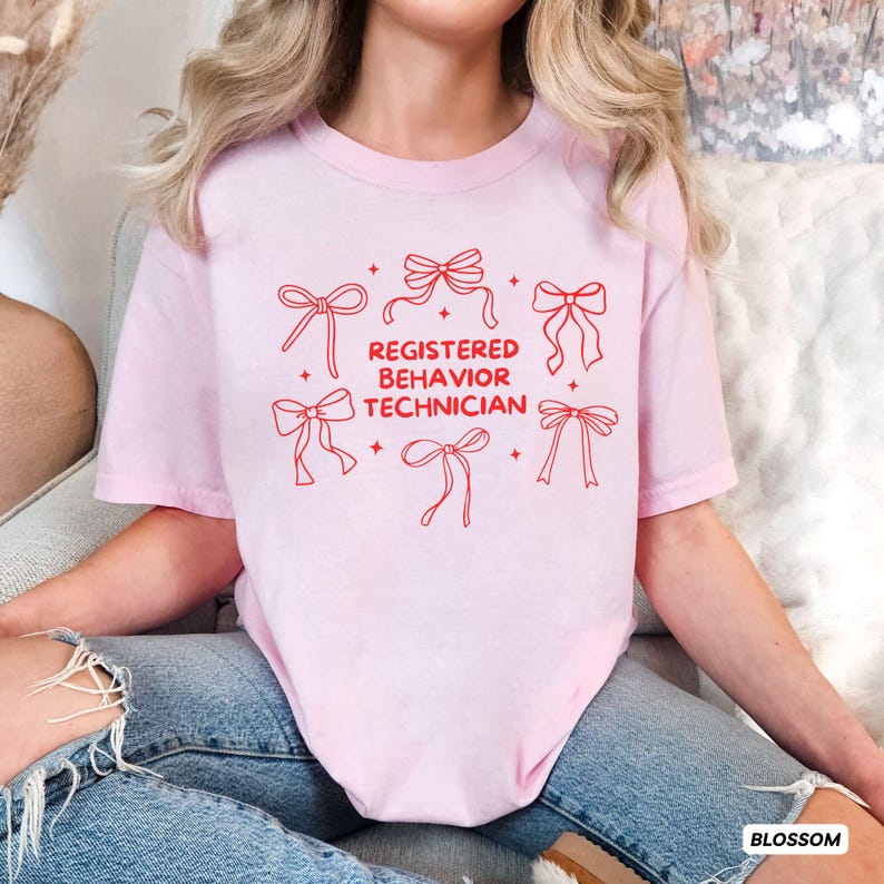 May include: A light pink short-sleeved t-shirt featuring red line art graphics of six bows arranged around the centred red text that reads REGISTERED BEHAVIOR TECHNICIAN, accented by small four-pointed star shapes scattered throughout the design.