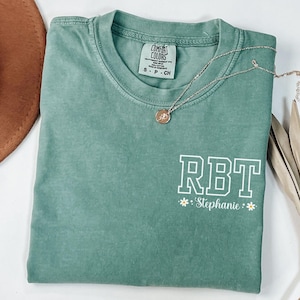 May include: A sage green Comfort Colors t-shirt with the letters "RBT" in white block letters, and the name "Stephanie" below. A silver necklace with a small pendant is draped across the shirt.