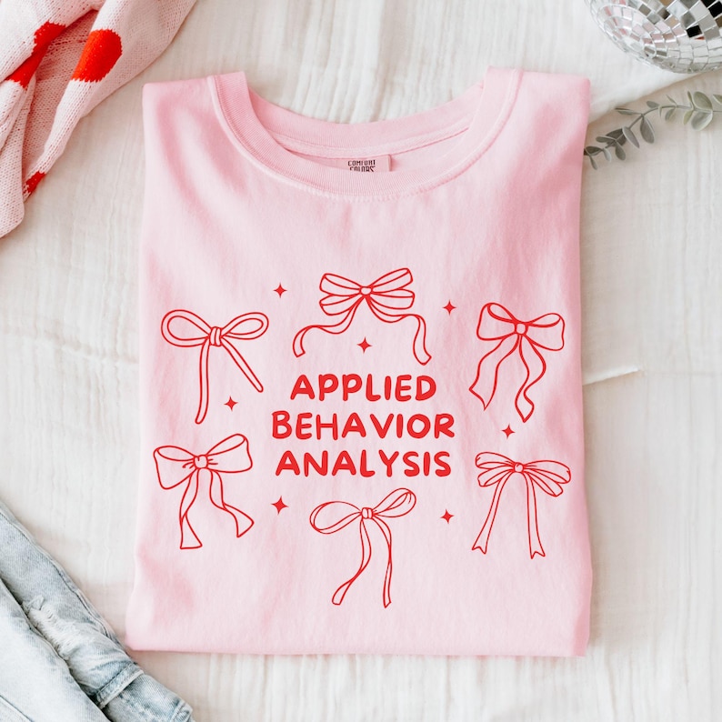 May include: Pink t-shirt with the text "APPLIED BEHAVIOUR ANALYSIS" in red lettering. The text is surrounded by red bows with stars.