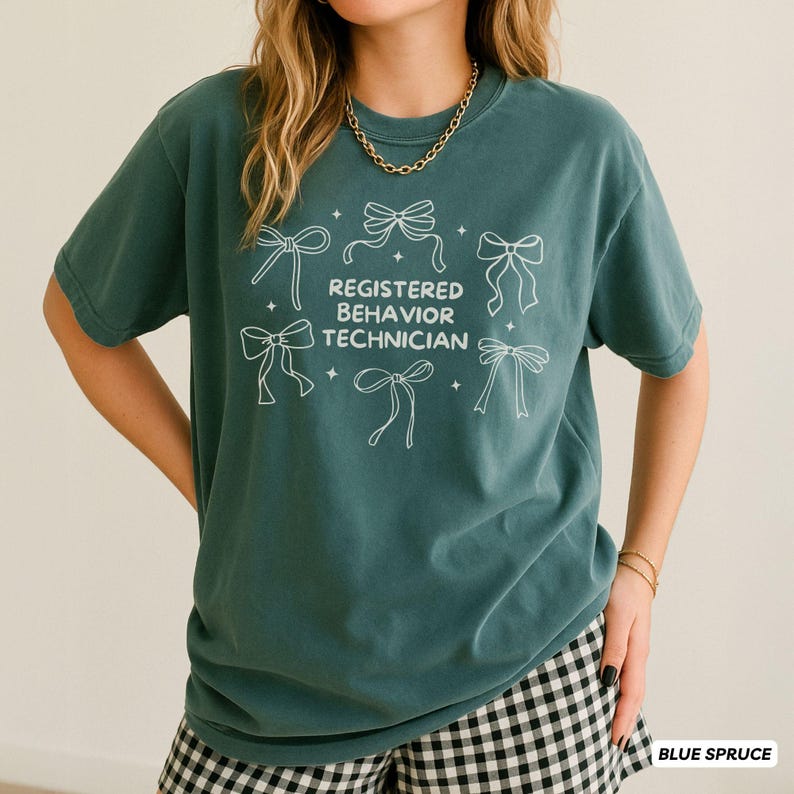 May include: A blue spruce coloured t-shirt featuring a white graphic print of five bows and the text Registered Behavior Technician centred on the front. The shirt has a crew neckline and short sleeves, providing a casual and comfortable fit.