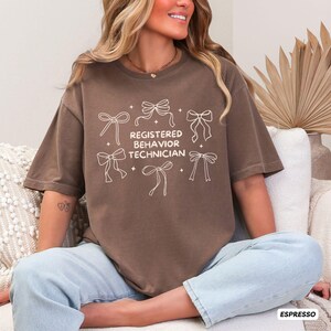 May include: A brown short-sleeve t-shirt featuring a cream-coloured graphic of six line-art bows arranged around the text "REGISTERED BEHAVIOR TECHNICIAN" with small star accents. The shirt has a crew neckline and a relaxed, loose-fitting cut.