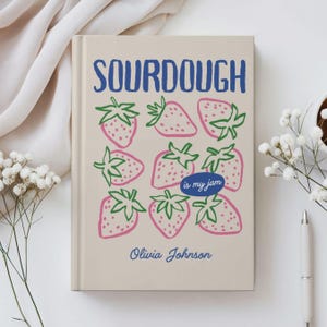 Puede incluir: Un diario beige con la palabra "SOURDOUGH" en azul, sobre un patr&oacute;n de fresas rosas con tallos verdes. El texto "is my jam" est&aacute; en un &oacute;valo azul, y el nombre "Olivia Johnson" est&aacute; debajo.