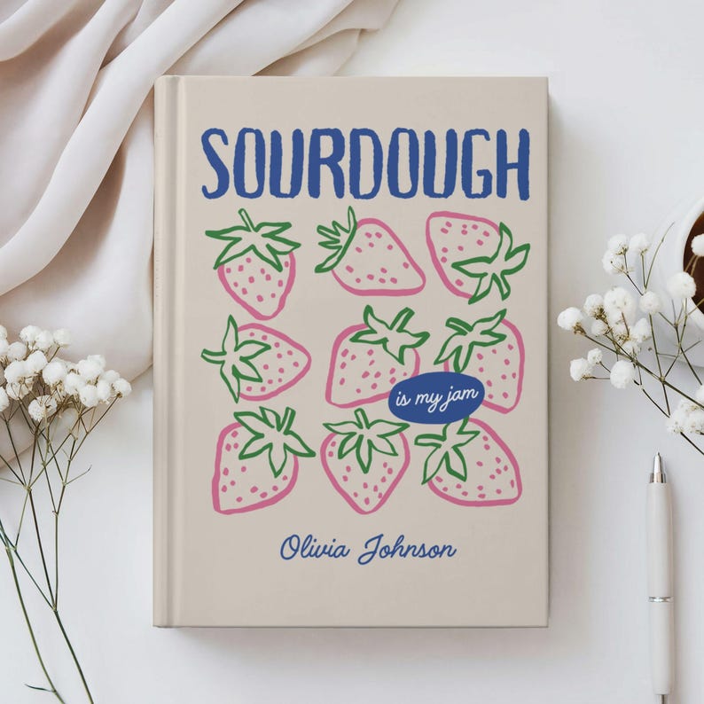 Puede incluir: Un diario beige con la palabra "SOURDOUGH" en azul, sobre un patr&oacute;n de fresas rosas con tallos verdes. El texto "is my jam" est&aacute; en un &oacute;valo azul, y el nombre "Olivia Johnson" est&aacute; debajo.
