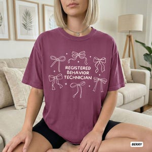 May include: A berry-coloured short-sleeve t-shirt featuring a white graphic print of six decorative bows and small stars surrounding the text "REGISTERED BEHAVIOR TECHNICIAN" in a clean, sans-serif font centred on the chest area.