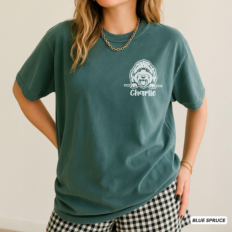 Op de afbeelding: Een blauwspar-kleurig t-shirt met korte mouwen en een witte afbeelding van een hondengezicht op de linkerborst. Onder de hondenillustratie staat de naam Charlie gedrukt in een wit cursief lettertype. Het shirt heeft een ronde hals.