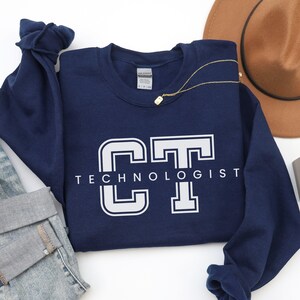 May include: A navy blue crewneck sweatshirt featuring the white collegiate-style letters CT with the word TECHNOLOGIST printed in white underneath. The garment has long sleeves and a Gildan brand label visible at the inner neckline.