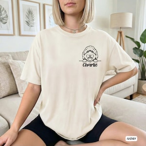Op de afbeelding: Een ivoorkleurig T-shirt met korte mouwen en een zwarte lijntekening van een hondenhoofd dat boven een horizontale lijn uitsteekt, met de naam Charlie in een cursief lettertype direct onder de hondenillustratie op de borst gedrukt.