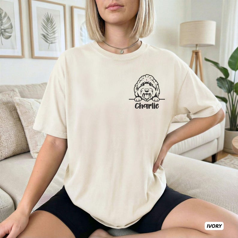 Op de afbeelding: Een ivoorkleurig T-shirt met korte mouwen en een zwarte lijntekening van een hondenhoofd dat boven een horizontale lijn uitsteekt, met de naam Charlie in een cursief lettertype direct onder de hondenillustratie op de borst gedrukt.