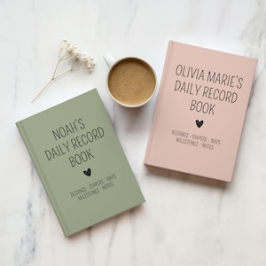 May include: Two hardcover daily record books, one sage green labeled Noah's Daily Record Book and one dusty pink labeled Olivia Marie's Daily Record Book, both featuring black text for feedings, diapers, naps, milestones, and notes.