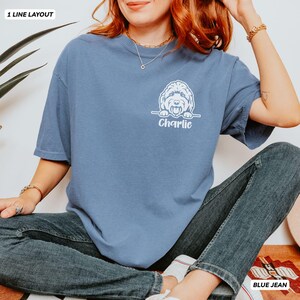 Op de afbeelding: Een blauw t-shirt met korte mouwen en een witte afbeelding van een hond die over een lijn kijkt, met de naam Charlie eronder gedrukt in een wit scriptlettertype. Het shirt heeft een ronde hals en een relaxte pasvorm, gecombineerd met jeans.