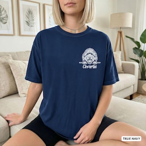 Op de afbeelding: Een marineblauw t-shirt met korte mouwen en een witte afbeelding van een hondengezicht dat boven een horizontale lijn uitsteekt, met de naam Charlie eronder gedrukt in een strak schreefloos lettertype op de linkerborst.