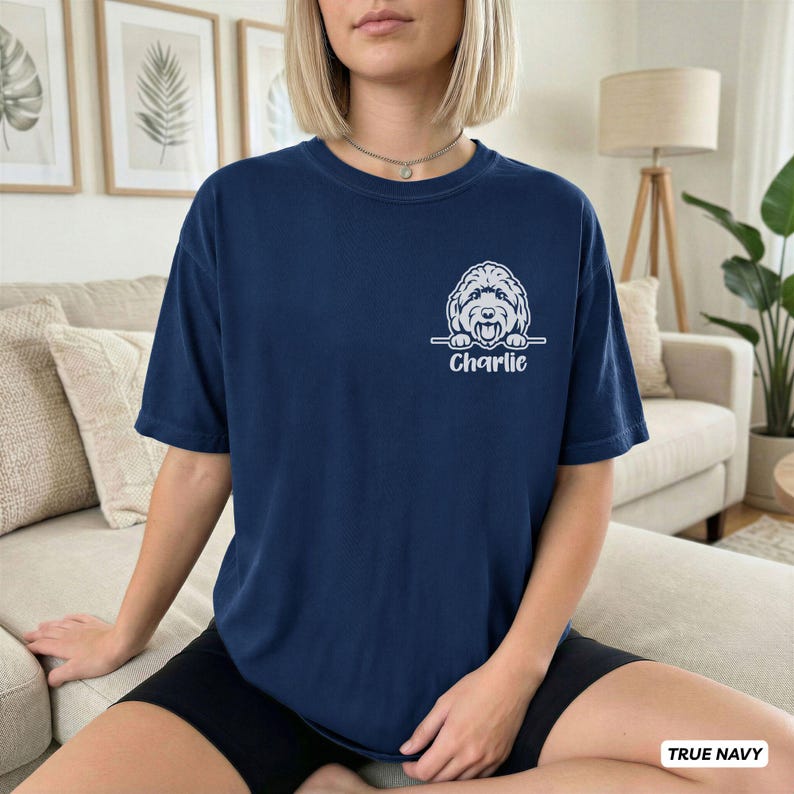 Op de afbeelding: Een marineblauw t-shirt met korte mouwen en een witte afbeelding van een hondengezicht dat boven een horizontale lijn uitsteekt, met de naam Charlie eronder gedrukt in een strak schreefloos lettertype op de linkerborst.