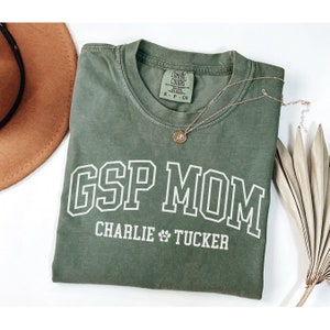 May include: Green t-shirt with white text that reads "GSP MOM CHARLIE TUCKER" with a paw print between the names.
