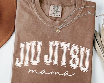 May include: A brown t-shirt with the words "Jiu Jitsu mama" in white lettering. The shirt has a crew neck and a small tag at the neckline. A silver necklace with a small pendant is draped across the shirt.