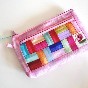 May include: Pink rectangular pouch with a colorful patchwork design. The patchwork features vertical and horizontal strips of various colors, including red, blue, orange, and purple. A small embroidered flower is on the corner. The pouch has a zipper closure and a tassel.