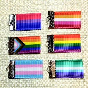 May include: Six ribbon keyrings with LGBTQ+ pride flag designs. The keyrings have a metal clasp and a ribbon with colourful stripes representing different pride flags, including rainbow, transgender, and bisexual flags.