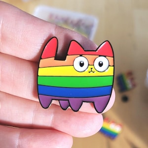 May include: A rainbow-striped cat enamel pin. The pin has a cartoon-like design with a red head and ears, and a body made up of horizontal stripes in rainbow colours. The cat has large, round eyes and is held in a hand.