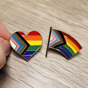 May include: Two enamel pins with gold borders. One is heart-shaped, the other a flag. Both feature the Progress Pride flag design, with rainbow stripes and additional colours representing inclusivity.