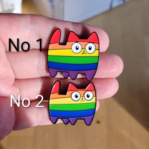 May include: Two enamel pins shaped like cats with rainbow-striped bodies, red ears, and purple legs. The top pin is labelled No 1 and the bottom pin is labelled No 2. Each cat has large white eyes, a small nose, and a mouth, outlined in black metal.
