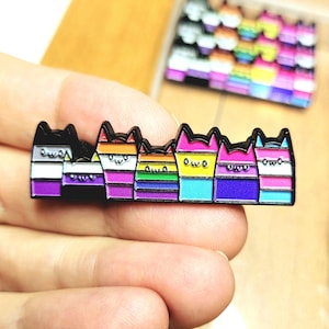 May include: A rectangular enamel pin featuring six stylised cat heads arranged in a row. Each cat has a different colour pattern representing various pride flags, with simple black outlines and a small owo face expression on each cat head.