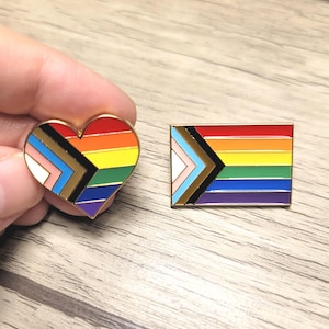 May include: Two enamel pins with gold borders. One is heart-shaped, the other rectangular. Both feature the Progress Pride flag design, with rainbow stripes and additional colours representing inclusivity.