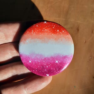 May include: A round button featuring a pink, white and orange ombre design. The button has a glittery texture with white stars scattered across the surface.