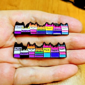 May include: Two black cat-shaped enamel pins with rainbow pride flag designs. Each cat has a different flag design, including the non-binary, lesbian, and pansexual flags. The pins have the word "owo" on them.