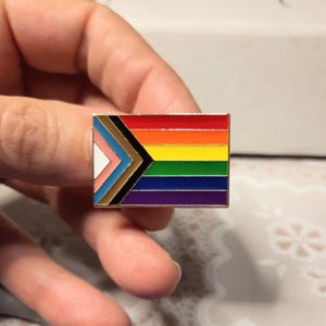 May include: A gold-coloured enamel pin featuring a rainbow flag with a black, brown, light blue, pink, and white chevron design in the top left corner.