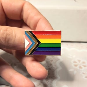 May include: A rectangular enamel pin featuring the Progress Pride flag design. The pin displays horizontal stripes in red, orange, yellow, green, blue, and violet, with a chevron on the left side containing white, pink, light blue, brown, and black stripes.