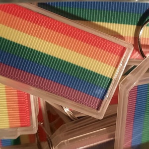 May include: A close-up of a stack of rainbow-coloured keyrings. Each keyring features a rainbow-striped ribbon with a clear plastic frame and a silver key ring.