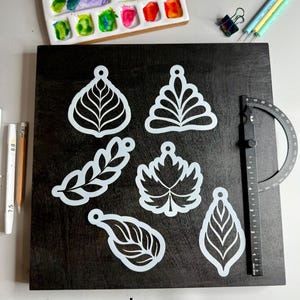 May include: White leaf-shaped stencils on a dark wooden surface, with a watercolor palette and art tools in the background. The word "Large" is printed at the bottom. The stencils include various leaf designs.